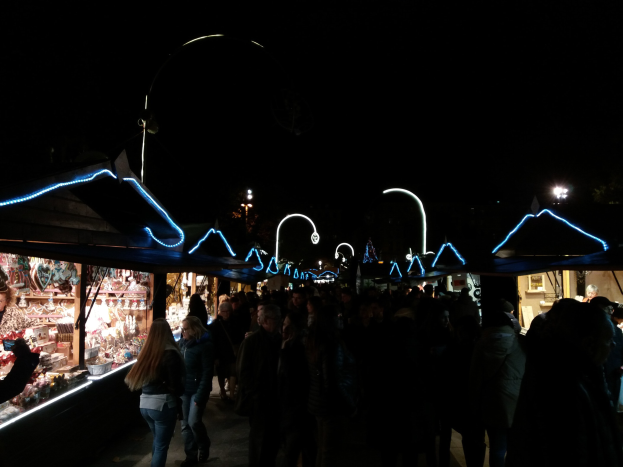 Eine Menschenmenge, die nachts eine Straße entlanggeht, gesäumt von beleuchteten Ständen mit verschiedenen Artikeln, mit einem dunklen Hintergrund, der die festliche Atmosphäre betont.