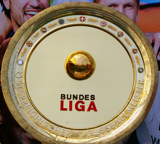 Ein Mann hält ein goldenes Schild mit 'Bundesliga' darauf hoch, im Hintergrund eine Fahne mit drei Personen.