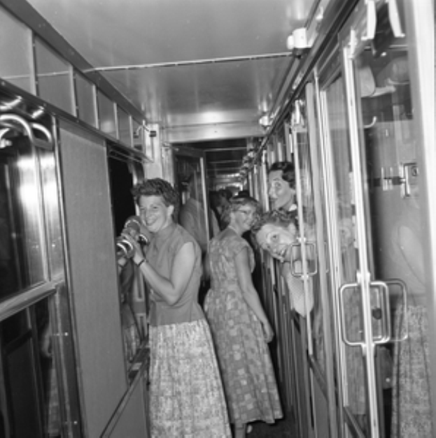 Eine Gruppe von Frauen, die auf einem Zug mit Türen auf beiden Seiten und Lampen oben stehen, dargestellt in Schwarz-Weiß.