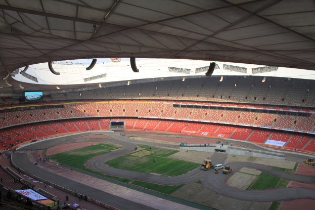 Großes Stadion voller Zuschauer, Fahrzeuge auf der Straße, grüner Boden, Texttafeln, Geländer, ein Bildschirm und Deckenbeleuchtung.
