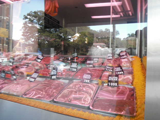 Eine Glasvitrine in einem Fleischgeschäft, die Tabletts mit Fleisch und Preisschildern zeigt, mit Gebäuden und Bäumen durch die Scheibe und einer Decke oben.