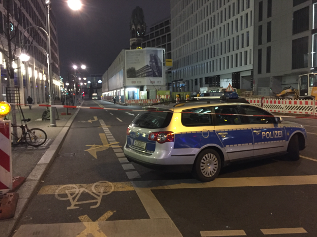 Ein Polizeiwagen parkt nachts an der Straße in Berlin, umgeben von Gebäuden, Straßnpfählen, Straßenlaternen, Kraftfahrzeugen, Barrieren, Fahrrädern, Personen auf dem Gehweg, Werbetafeln und einer Statue im Hintergrund.