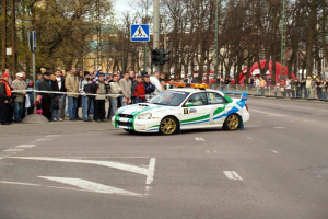 Ein Rallyefahrzeug fährt an Zuschauern vorbei auf einer Straße mit Bäumen, Masten, Verkehrszeichen und Gelöndern, mit Gebäuden und einem klaren Himmel im Hintergrund.