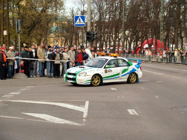 Ein Rallyefahrzeug fährt an Zuschauern vorbei auf einer Straße mit Bäumen, Masten, Verkehrszeichen und Gelöndern, mit Gebäuden und einem klaren Himmel im Hintergrund.