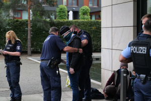 Eine Gruppe von Polizisten steht vor einem Gebäude zusammen, eine Frau hält ein Buch und einen Stift in der Hand, während mehrere Menschen in der Nähe stehen und sitzen und im Hintergrund Pflanzen, Bäume und Gebäude zu sehen sind.