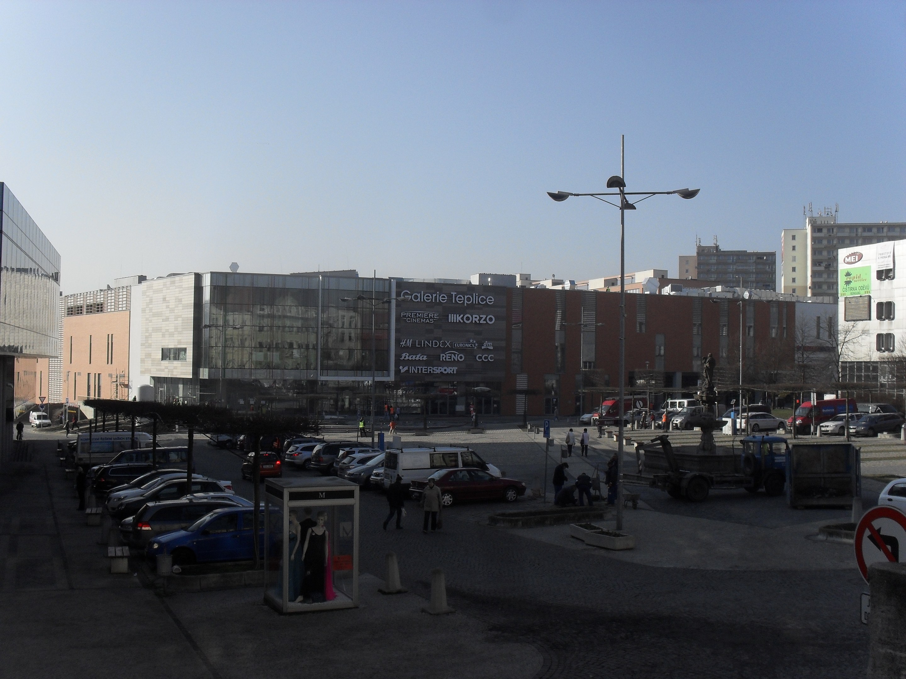 Eine belebte Stadtstraße mit parkenden Autos auf einem Parkplatz neben hohen Gebäuden, beleuchtet von Straßenlaternen, mit einigen Fußgängern und dem Himmel im Hintergrund.