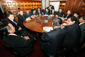Eine Runde-Tisch-Besprechung mit Delegierten, die über Mikrofone, Papiere, Kabel und Dateien auf dem Tisch sprechen, mit einer hölzernen Wand im Hintergrund.