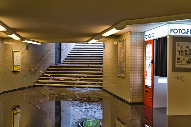 Eine überflutete U-Bahn-Station in Berlin mit Wasser auf dem Boden, Treppen mit Geländern, gerahmten Fotos an den Wänden, Deckenleuchten und einem Getränkeautomaten.