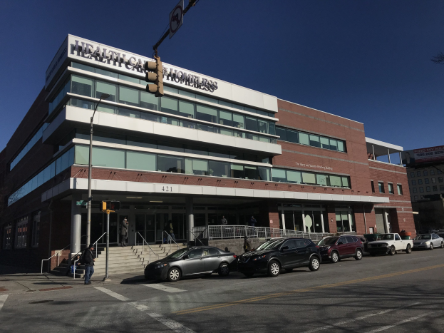 Großes Gebäude, das als New York City Health Care Homeless Center bezeichnet wird, mit geparkten Autos, Fußgängern, Straßenmöbeln und Bäumen unter einem klaren Himmel.