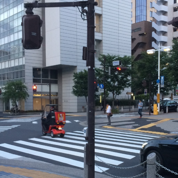 Scooter auf einer Straße mit Menschen im Hintergrund, einer Ampel im Vordergrund und Gebäuden mit Bäumen im Hintergrund.