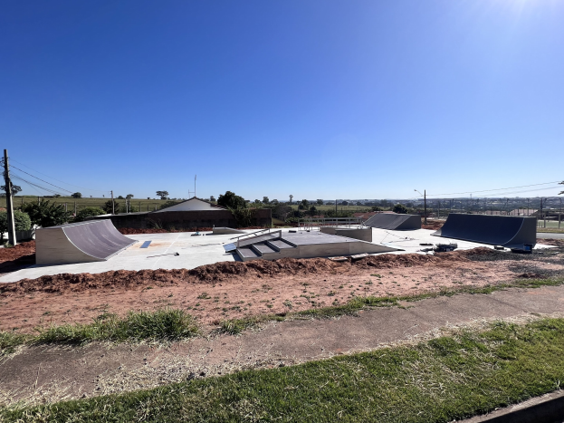Ein Skatepark mit mehreren Rampen, umgeben von Gras und Bäumen, Pfählen und Drähten im Hintergrund und einem klaren blauen Himmel darüber.