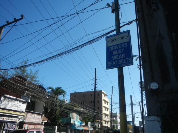 Eine Stra√e in einer Stadt mit Autos, Stromm√§sten mit Kabeln, Geb√§uden, B√§umen und Schilden, auf denen ein Schild an einem Strommast zu sehen ist, auf dem steht: "F√ľhrende m√ľssen den Sicherheitsgurt tragen."