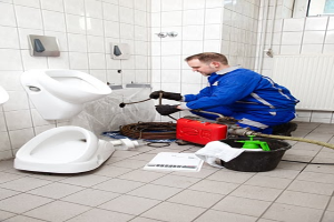 Ein Mann in einer blauen Uniform sitzt auf dem Boden vor einem Urinal und hält eine Pfeife in der Hand, umgeben von Reinigungsmitteln, mit Urinalen an der linken Wand und einem Fenster auf der rechten Seite.