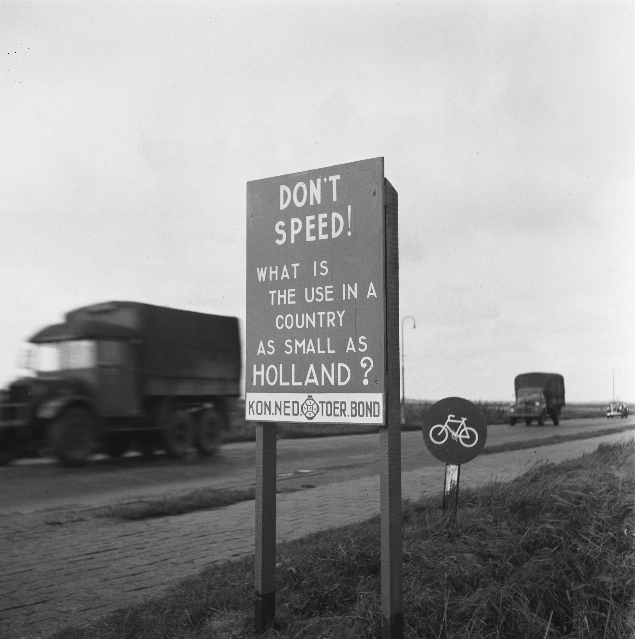 Schwarz-weißes Foto eines Lkws auf einer Straße mit einem "Fahr nicht zu schnell"-Schild im Vordergrund und anderen Fahrzeugen, Gras, Pfosten und einem klaren Himmel im Hintergrund.