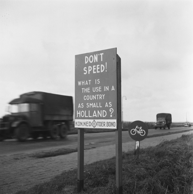 Schwarz-weißes Foto eines Lkws auf einer Straße mit einem "Fahr nicht zu schnell"-Schild im Vordergrund und anderen Fahrzeugen, Gras, Pfosten und einem klaren Himmel im Hintergrund.