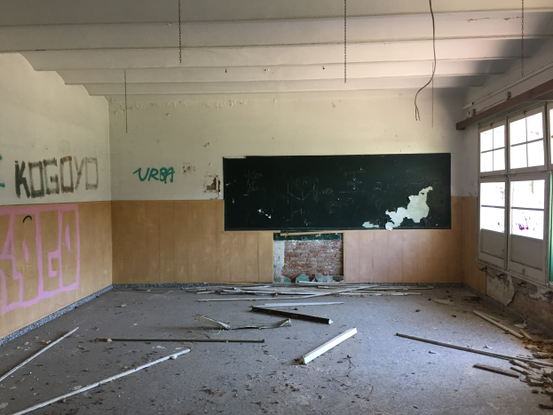 Leerer Klassenraum mit Graffiti an den Wänden, einer zentralen Tafel, Glasfenstern auf der rechten Seite und einem Boden, der mit zerbrochenen Holzstücken übersät ist.