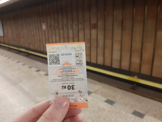 Eine Person, die eine Fahrkarte in einer U-Bahn-Station hochhölt, mit einem Bahnsteig auf der linken Seite und einer Wand im Hintergrund.