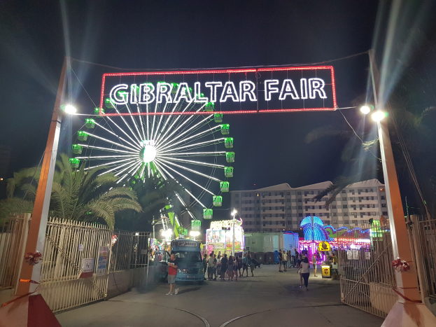 Der Eingang zur Gibraltar-Messe bei Nacht, beleuchtet von einem großen Riesenrad und einer leuchtenden Tafel, mit Menschenmassen, Bäumen und Gebäuden im Hintergrund unter einem dunklen Himmel.
