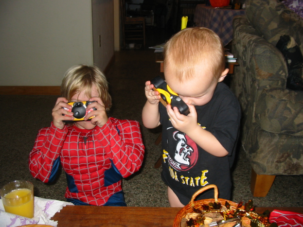 Zwei Kinder, eines im Spider-Man-Kostüm, halten eine Spielzeugkamera in der Nähe eines Holztisches mit einem Glas und einem Korb, mit einer Couch auf der rechten Seite und einem weiteren Holztisch im Hintergrund.