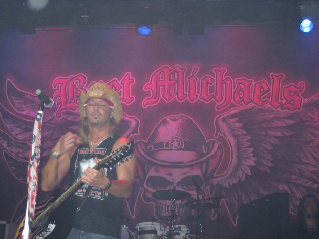 Eine Musik-Konzertszene mit Scheinwerfern an der Spitze, einem Banner im Hintergrund, Drogen in der Mitte unten, einer Person in der rechten unteren Ecke sitzen und einem Gitarristen, der vor einem Mikrofon auf der linken Seite spielt.