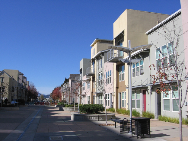 Stadtstraße mit Wohngebäuden, Bäumen, Laternen, Bänken und Mülltonnen gesäumt, mit Hügeln und einem klaren blauen Himmel im Hintergrund.