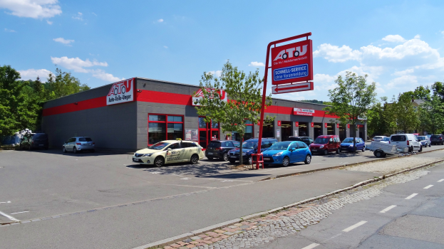 Ein Autohaus mit parkenden Fahrzeugen vorne, umgeben von Gebäuden, Bäumen, Strommasten, Schildern und einer Straße unter einem bewölkten Himmel.