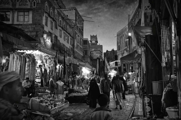 A black and white image of a bustling market scene with people walking and standing, shops with hanging dresses, buildings with windows, a bucket, and a name board, with a watermark in the top right corner.