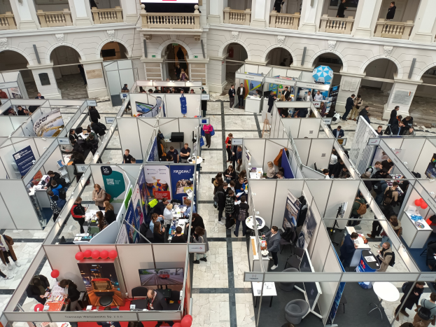 Große Gruppe von Menschen auf einer internationalen Messe mit Ständen, Bannern, Tischen, Stühlen und Ballons in einem Raum mit gewölbter Architektur und einem Bildschirm im Hintergrund.
