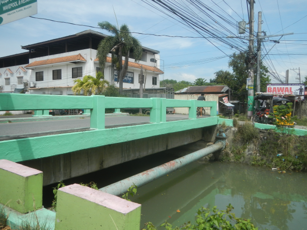 Eine grüne Brücke mit Geländern überspannt einen kleinen städtischen Bach, umgeben von Pflanzen und Wasser, mit Strommasten, Texttafeln und Fahrzeugen in der Nähe und Gebäuden, Bäumen und bewölktem Himmel im Hintergrund.