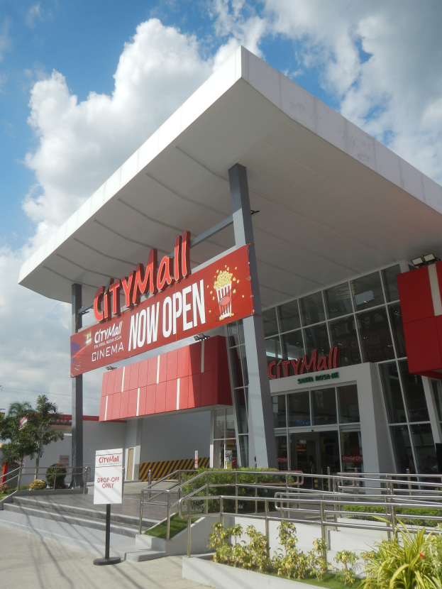 Ein belebter Stadtmarkt mit einem Schild "City Mall Now Open" umgeben von Gebäuden, Namensschildern, Geländern, Pflanzen, Bäumen, Strommasten mit Drähten und einem bewölkten Himmel im Hintergrund.