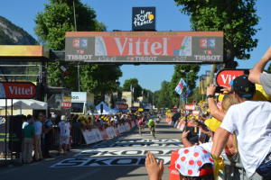 Eine Gruppe von Radfahrern fährt auf einer Straße während der Tour de France 2016, mit Zuschauern an den Seiten, Bäumen, einem Hügel und einem klaren blauen Himmel im Hintergrund und einem Bogen mit "Tour de France 2016" oben.
