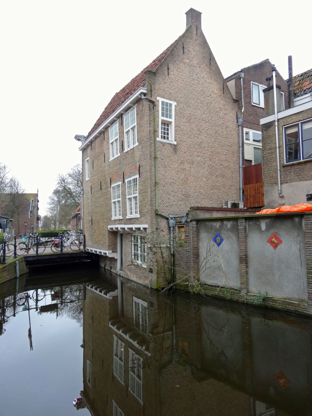 Kanal in Delft, Niederlande, mit einem Gebäude auf der rechten Seite, Fahrrädern auf der linken Seite, Bäumen im Hintergrund und einem klaren blauen Himmel darüber.