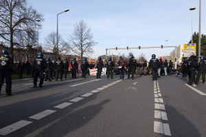 Gruppe von Polizisten in schwarzen Uniformen und Helmen, die an der Seite einer Straße mit Laternen, Ampeln, Bäumen, Gebäuden und einem klaren blauen Himmel im Hintergrund stehen.