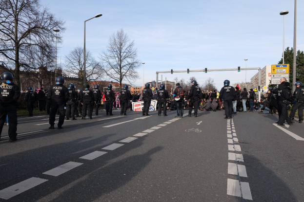 Gruppe von Polizisten in schwarzen Uniformen und Helmen, die an der Seite einer Straße mit Laternen, Ampeln, Bäumen, Gebäuden und einem klaren blauen Himmel im Hintergrund stehen.