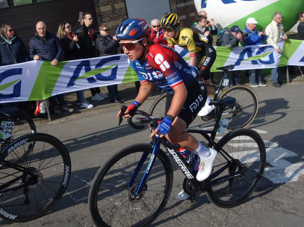 Eine Gruppe von Radfahrern, die während der Tour de France eine Straße hinunterfahren, während Zuschauer hinter ihnen Schilder halten und ein Gebäude mit Fenstern im Hintergrund zu sehen ist.