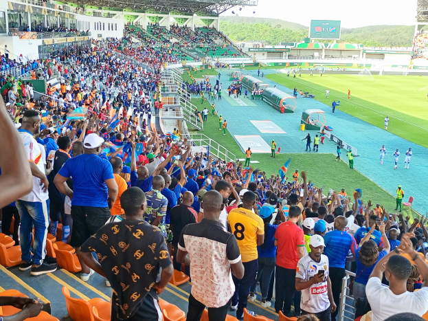 Eine große Menschenmenge versammelt sich vor einem eingezäunten Fußballfeld mit Hügeln und einem klaren blauen Himmel und wartet gespannt auf den Beginn eines Spiels.
