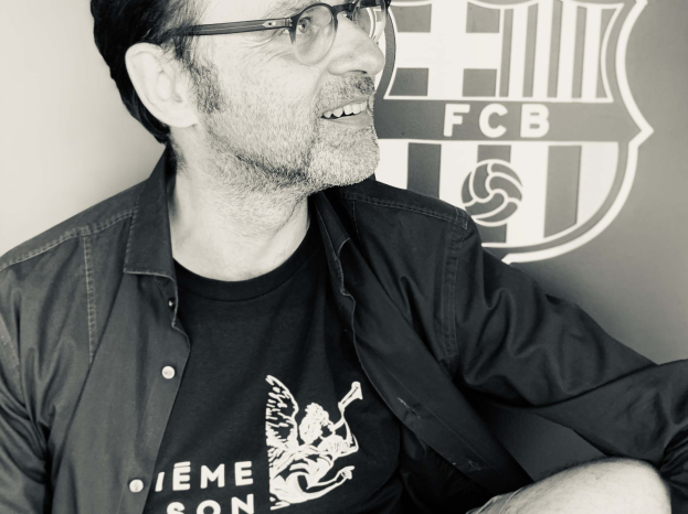 Ein Mann in einer Brille und einem schwarzen Hemd lächelt, während er vor dem FC Barcelona-Logo an einer Wand sitzt, auf einem Schwarz-Weiß-Bild.