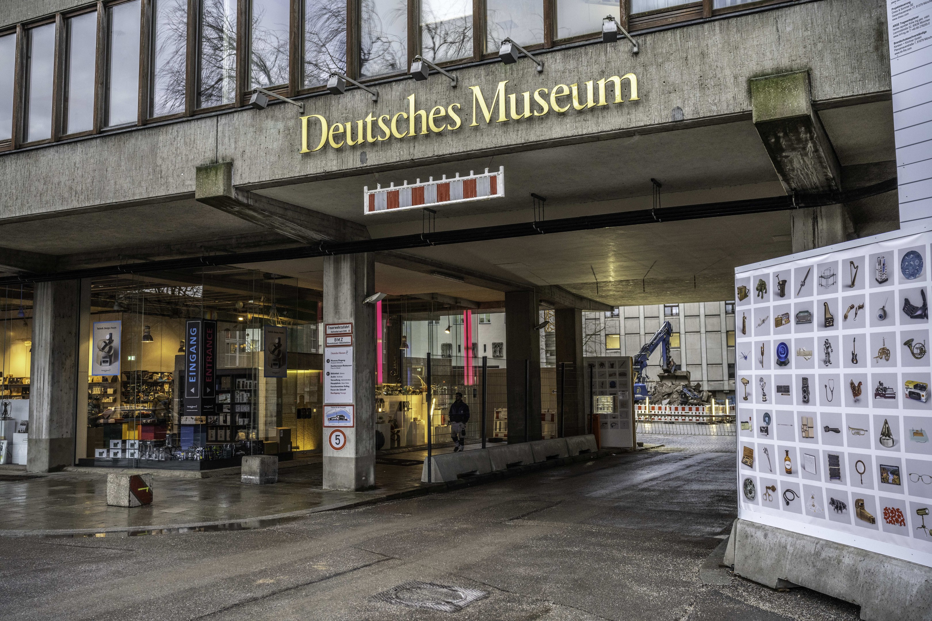 Au\ssenseite des Deutschen Museums in Berlin, mit einem gro\ssen Geb"ude mit Glasfenstern, S"ulen und einer Namensplakette, neben einer Informationsplakette, mit B"umen, anderen Geb"uden und dem Himmel im Hintergrund.