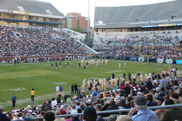 Fußballspiel in einem Stadion mit Spielern auf dem Feld und Zuschauern in den Rängen unter einem blauen Himmel.