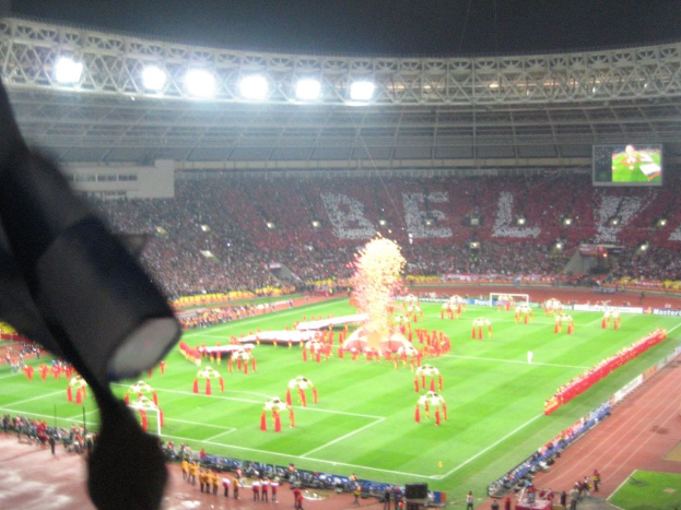 Ein Stadion mit Flutlicht, Eisengittern, Zuschauern, stehenden Individuen, Werbetafeln, Anzeigetafeln, Sicherheitskameras, Flaggen und Décor.