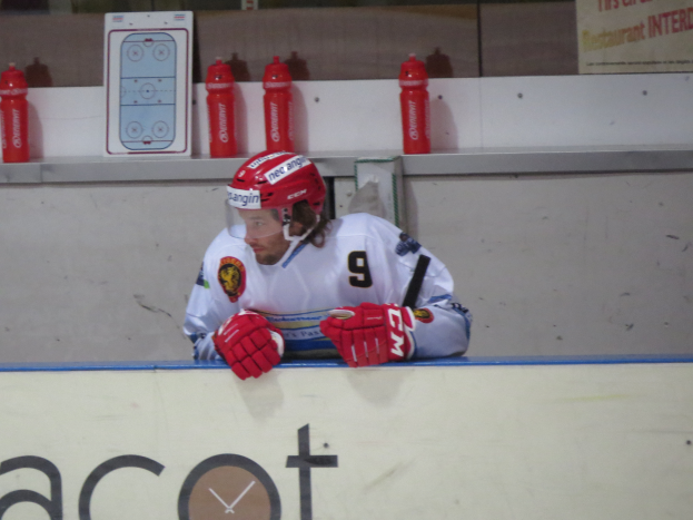 Ein Hockey-Spieler sitzt auf einer Bank und trägt einen Helm und Handschuhe, mit Flaschen und einer Tafel mit Schriftzug an der Wand dahinter.