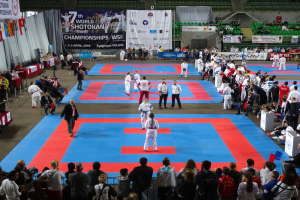 Eine Gruppe von Menschen steht auf einer blauen und roten Judo-Matte, umgeben von einer Menge, mit Tischen, Bannern und Flaggen im Hintergrund bei den 7. World Shotokan Championships in Polen.