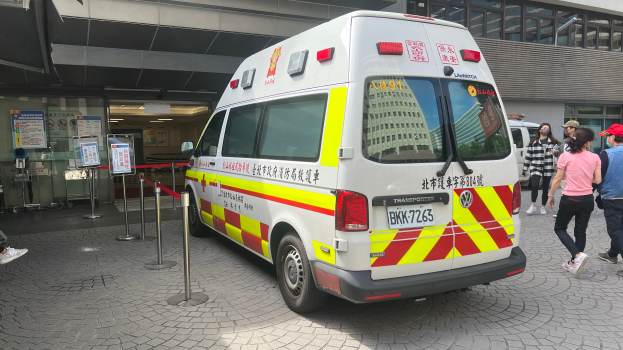 Ein Ambulanzfahrzeug steht vor einem Gebäude mit Menschen auf der rechten Seite, eine Person sitzt auf einem Stuhl im Hintergrund und Schilder mit Text und Plakaten an der linken Wand, auf denen «Ambulance Service in Hong Kong» steht.