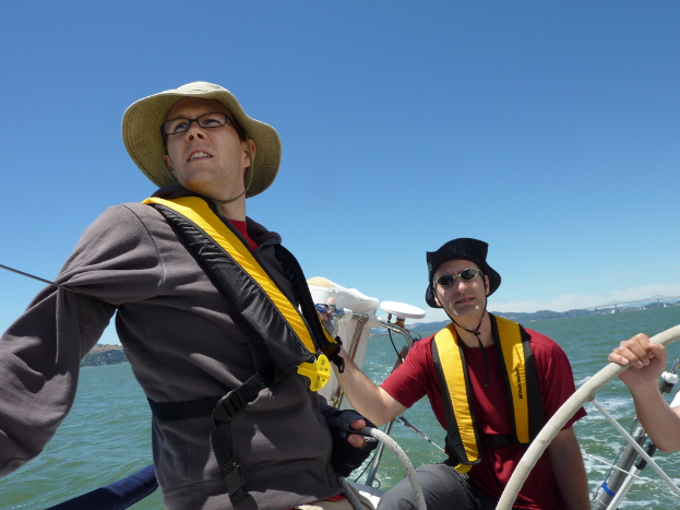 Zwei Männer auf einem Boot, einer trägt einen braunen Hut und ein graues Kleidungsstück, der andere einen schwarzen Hut und ein rotes T-Shirt, mit dem Meer im Hintergrund.