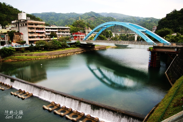 Eine Brücke über einen Fluss mit Gebäuden, Bäumen, Boden, Hügeln, Zäunen und Himmel im Hintergrund.