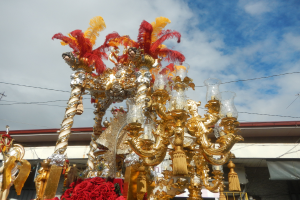 Ein großes goldenes und rotes Festwagen, geschmückt mit Blumen und anderen Dekorationen bei einem Karnevalsumzug, mit einem Gebäude, Strommasten mit Drähten und einem bewölkten Himmel im Hintergrund.
