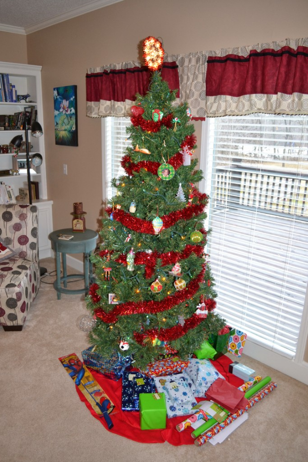 Ein Weihnachtsbaum mit Geschenken vorne, eine Couch, ein Tisch, ein Bücherregal, ein wandhängendes Bild und eine Stehlampe mit Vorhängen im Hintergrund.