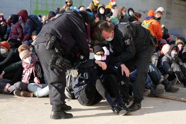 Eine Gruppe von Menschen, einige mit Mützen und Masken, sitzt auf dem Boden, während zwei Polizeibeamte in der Nähe stehen, in einer städtischen Umgebung mit einer Wand im Hintergrund.