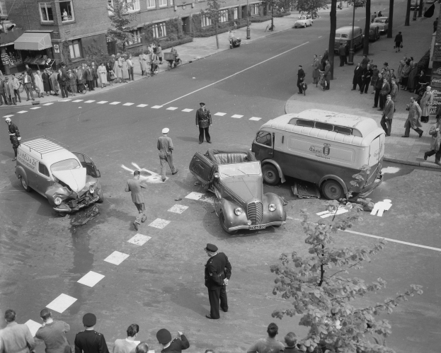 Schwarz-weißes Bild einer Autounfall-Szene an einer Straßenkreuzung mit mehreren Fahrzeugen, darunter ein Van, und einer Gruppe von Zuschauern; Gebäude, Bäume, Pfosten und Texttafeln sind im Hintergrund zu sehen.