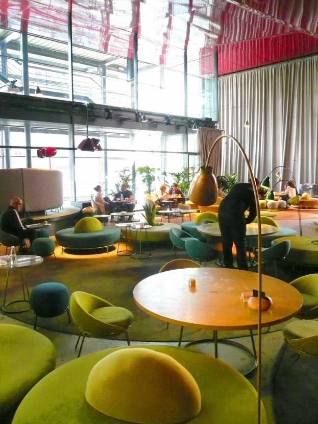 Ein geräumiges Hotel-Lounge mit grünen Stühlen, Tischen, Sofas, stehenden Menschen, Pflanzen, einer Stehlampe, Vorhängen und Deckenleuchten.